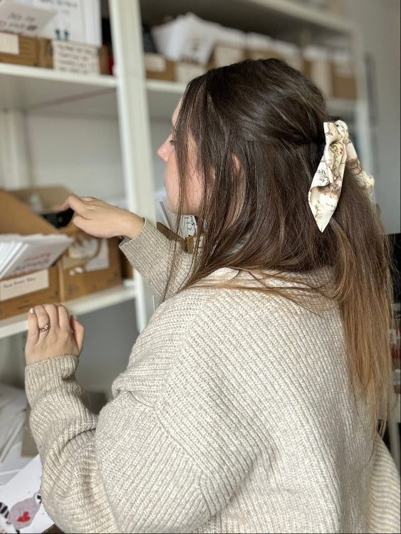 Abbie, the illustrator behind Abbie Imagine, is seen from behind, looking at and picking greeting cards on white shelves filled with boxes and greeting cards.